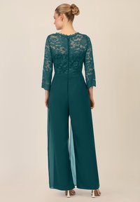 Jumpsuit in pizzo teal con maniche a tre quarti, pannelli inferiori trasparenti e zip nascosta sul retro. Texture liscia e design con gamba ampia.