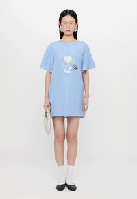 Helles blaues kurzes Kleid mit fließenden Ärmeln, gewelltem Saum und floralem Muster. Getragen mit weißen Socken und schwarzen Ballerinas, während eine beigefarbene Tasche gehalten wird.