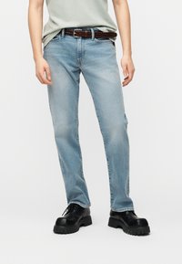 Άτομο που φοράει ανοιχτό μπλε τζιν με καφέ πλεκτή ζώνη και μαύρα χοντρά δερμάτινα παπούτσια με κορδόνια, στέκε ται μπροστά σε ένα απλό λευκό φόντο.