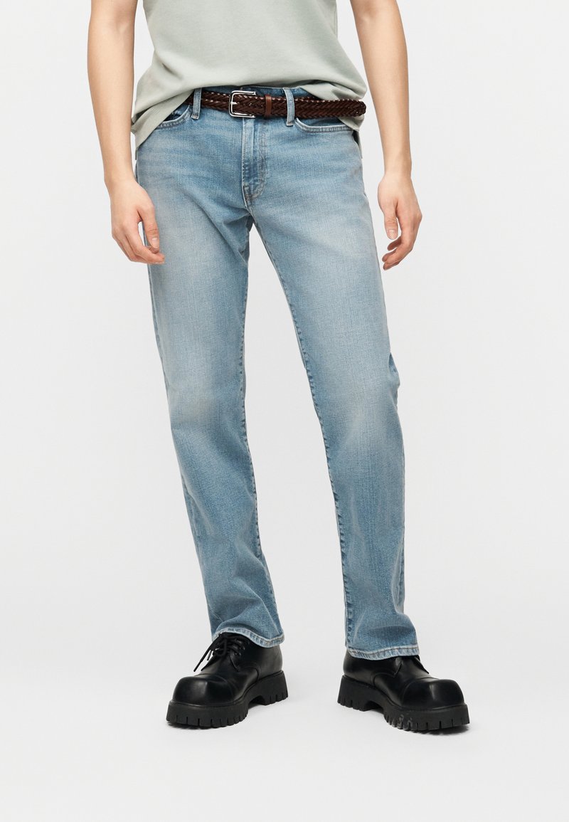 Άτομο που φοράει ανοιχτό μπλε τζιν με καφέ πλεκτή ζώνη και μαύρα χοντρά δερμάτινα παπούτσια με κορδόνια, στέκεται μπροστά σε ένα απλό λευκό φόντο.