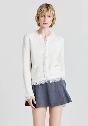 Veste blanche texturée avec des bords effilochés et deux poches avant, dotée de quincaillerie dorée. Associée à une jupe grise évasée.