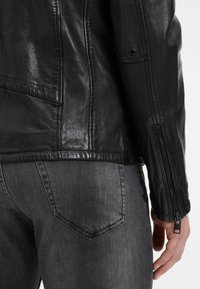 Giacca in pelle nera con finitura testurizzata, dettagli con zip sulle maniche e vestibilità sartoriale, abbinata a jeans grigi scuri.