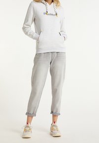 Žena v světle šedé mikině s žlutými akcenty, volných šedých vyhrnutých džínách a pestrých teniskách stojí s rukama v kapsách.