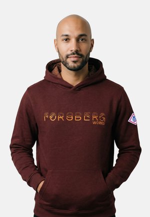 Mann mit Bart trägt einen dunkelroten Hoodie mit dem Schriftzug „Forsberg Works“ und steht mit den Händen in den Taschen vor einem einfarbigen weißen Hintergrund.