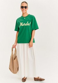 Zelené bavlnené tričko s nápisom "Merde!" napísaným žltou farbou, skombinované s bielou maxi sukňou, béžovou sieťovou taškou a topánkami s leopardím vzorom.
