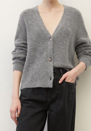 Persona con un cárdigan gris abotonado de mangas remangadas y pantalones negros, con una mano en el bolsillo, frente a un fondo claro y uniforme.