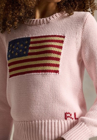 Femme portant un pull rose en maille avec un motif du drapeau américain et des initiales rouges "RL" près de l'ourlet.