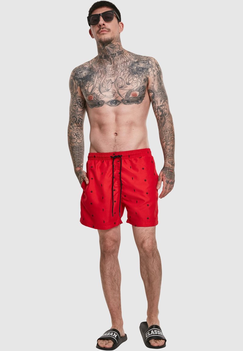 Hombre sin camiseta con tatuajes extensos lleva gafas de sol negras, pantalones cortos de baño rojos con patrón negro y sandalias negras con texto blanco sobre fondo gris.