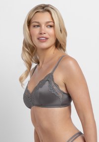 Blondinka, ki nosi sivi satenasti beli nedrček z čipko, rahlo obrnjena vstran, na preprostem svetlem ozadju.