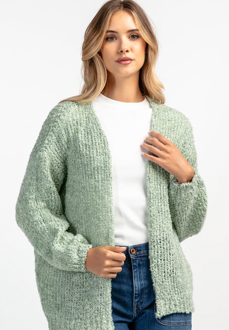 Jasnozielony, dziany kardigan o luźnym fasonie, z prążkowaną teksturą, otwartym przodem i opuszczonymi ramionami, noszony na białej bluzce.