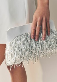 Weißes Clutch Bag mit einem strukturierten Perlenmuster am Boden, mit herabhängenden klaren und silbernen Perlen, in einer Hand gehalten.