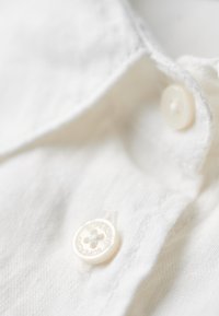 Biała lniana koszula zbliżenie na okrągły, biały guzik z tłoczonym tekstem. Tekstura tkaniny jest miękka, z widocznymi szwami.