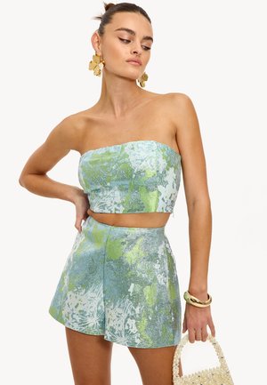 Femme portant un haut court sans bretelles à motif floral bleu et vert avec un short taille haute assorti, tenant un sac à main orné de perles.