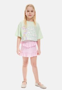 Overdimensioneret lys grøn t-shirt med grafik tekst, parret med en lyserød lagdelt flæse nederdel. Beige balletflade sko fuldender outfittet.