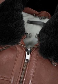 Braune Lederjacke mit schwarzem Schaffellkragen, ausgestattet mit einem silbernen Reißverschluss und strukturierter Verarbeitung. Das Innere zeigt ein hellfarbiges Futter.