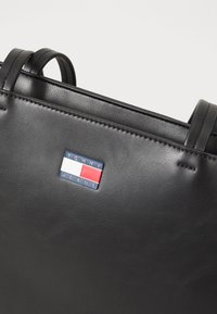Sac à main en cuir noir avec double poignée supérieure et forme rectangulaire. Présente un patch logo coloré avec du bleu, du blanc et du rouge. Surface texturée.
