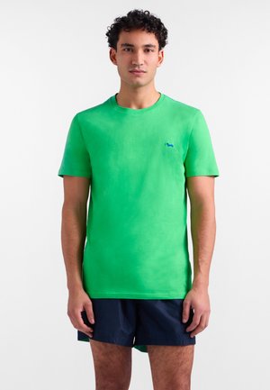 Mladý muž s krátkymi kučeravými vlasmi, oblečený v jasne zelenej krátkosleevej tričku a tmavomodrých šortkách na bielom pozadí.