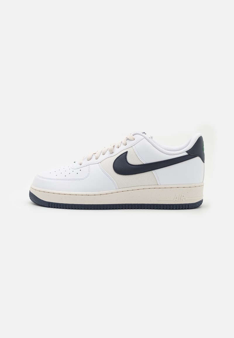 Baskets basses blanches Nike avec Swoosh noir, semelle intermédiaire beige, semelle extérieure noire et bout perforé sur fond blanc uni.
