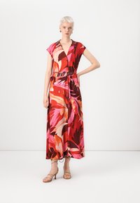 Robe maxi dans un motif floral vibrant rouge et rose, manches courtes, décolleté en V, taille ceinturée avec une boucle ronde, associée à des talons beiges à lanières.