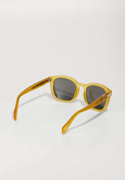 Óculos de sol de plástico amarelo com lentes cinzas. Montura arredondada com acabamento brilhante, apresentando hastes afiladas. Destacados com detalhes subtis nas dobradiças.
