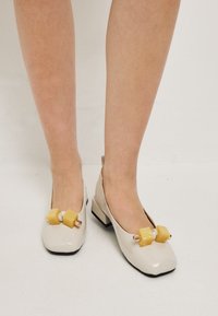 Ballerines en cuir verni beige avec un bout carré, des accents géométriques jaunes et un petit talon noir. Texture lisse avec des éléments décoratifs.