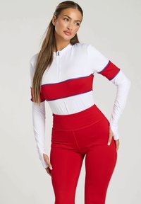 Tenue de sport rouge et blanche comprenant un haut zippé à manches longues avec une bande rouge horizontale et des leggings taille haute assortis. Texture lisse.