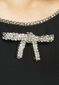 Gros plan d’un vêtement noir avec un col bordé de strass et une broche en forme de nœud ornée de strass épinglée sous le col.