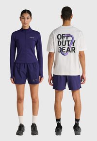 Frau steht vorn, trägt eine lila Jacke mit Reißverschluss und Shorts, Mann steht mit dem Rücken zur Kamera, trägt ein weißes T-Shirt mit dem Aufdruck "OFF DUTY GEAR" und lila Shorts.