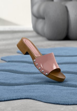 Sandale à enfiler rose avec des clous argentés et une semelle en bois sur un tapis bleu texturé avec un fond gris flou.