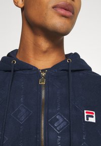 Felpa con zip blu navy, con zip dorata, motivo in rilievo con logo Fila e patch logo rosso/bianco. Tessuto testurizzato e cappuccio con cordino.
