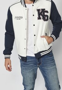 Veste varsity blanche et marine avec des poignets rayés, des boutons et un logo brodé. Elle présente une coupe décontractée et une texture de tissu douce.