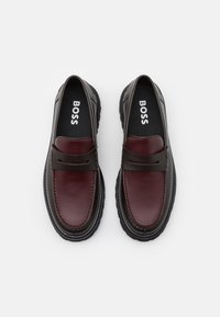 Mocassins en cuir de couleur marron foncé et bordeaux avec une finition texturée. Présentent une semelle en caoutchouc noir flexible et un design minimaliste.