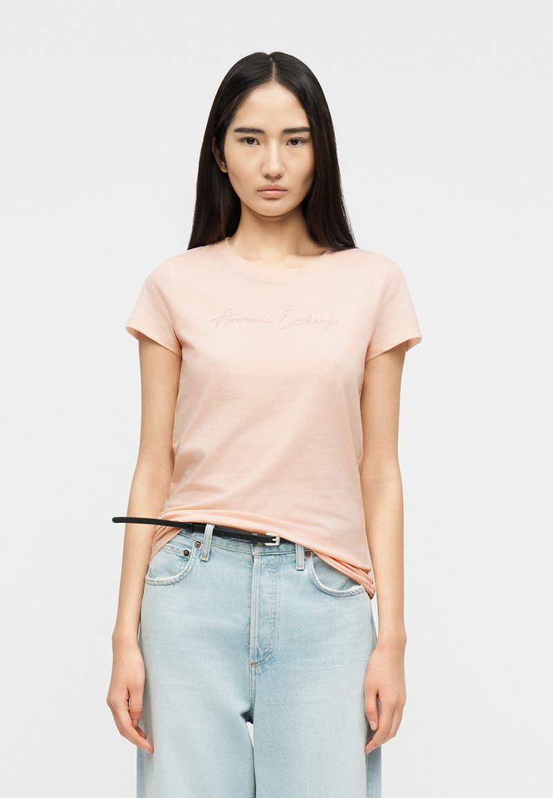 Νεαρή γυναίκα με μακριά σκούρα μαλλιά φοράει ροζ ανοιχτό κοντομάνικο μπλουζάκι και ανοιχτό μπλε τζιν ψηλόμεσο με μαύρη ζώνη, στέκεται ουδέτερα.