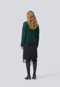 Femme avec de longs cheveux blonds ondulés portant un pull vert foncé, une jupe noire à dentelle, des collants noirs et des chaussures noires à petits talons, se tenant de dos.