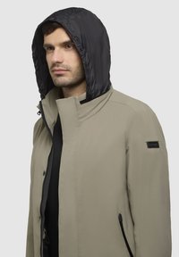 Beige jakke med sort hætte, lavet af glat stof; har ståkrave og sidelommer. Brandpatch på ærmet.