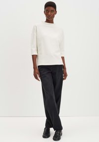 Donna in piedi con una maglietta bianca a maniche 3/4 texturizzata, pantaloni neri a gamba larga e scarpe nere senza lacci, su uno sfondo chiaro e semplice.
