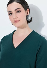 Turkusowa bluzka V-neck z teksturowanej tkaniny, z wyraźnym dekoltem. Duże, okrągłe szare kolczyki dopełniają projekt.
