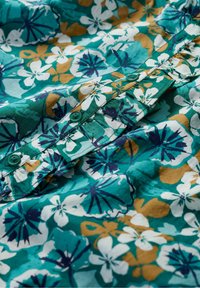 Bloemshirt met een teal achtergrond, witte en gele bloemen, en donkerblauwe accenten. Zachte stof met een knoopsluiting.