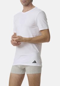 White short-sleeve t-shirt with a round neck and black logo accent on the lower left side, worn over light gray boxer shorts.