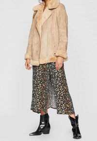 Donna che indossa un giubbotto in shearling beige, una gonna midi floreale e stivali neri in pelle alla caviglia, in piedi contro uno sfondo bianco.