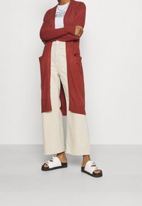 Röd lång cardigan med öppen framsida och fickor, kombinerad med krämfärgade vida byxor och vita sandaler med lager av remmar och svart sula.