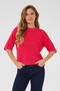 Hellrosa, gestrickter Pullover mit kurzen Ärmeln, Rundhalsausschnitt und strukturierter Oberfläche. Kombiniert mit dunkelblauen, enganliegenden Jeans.