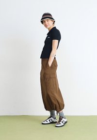 Camisa polo preta, calças cargo castanhas com bolsos, meias cinzentas e sapatilhas preto e brancas. Um chapéu com riscas completa o visual.