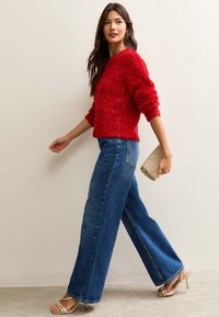 Czerwony, miękki sweter, szerokie niebieskie dżinsy z wysokim stanem, złote sandały na paski oraz mała metaliczna kopertówka na neutralnym tle.