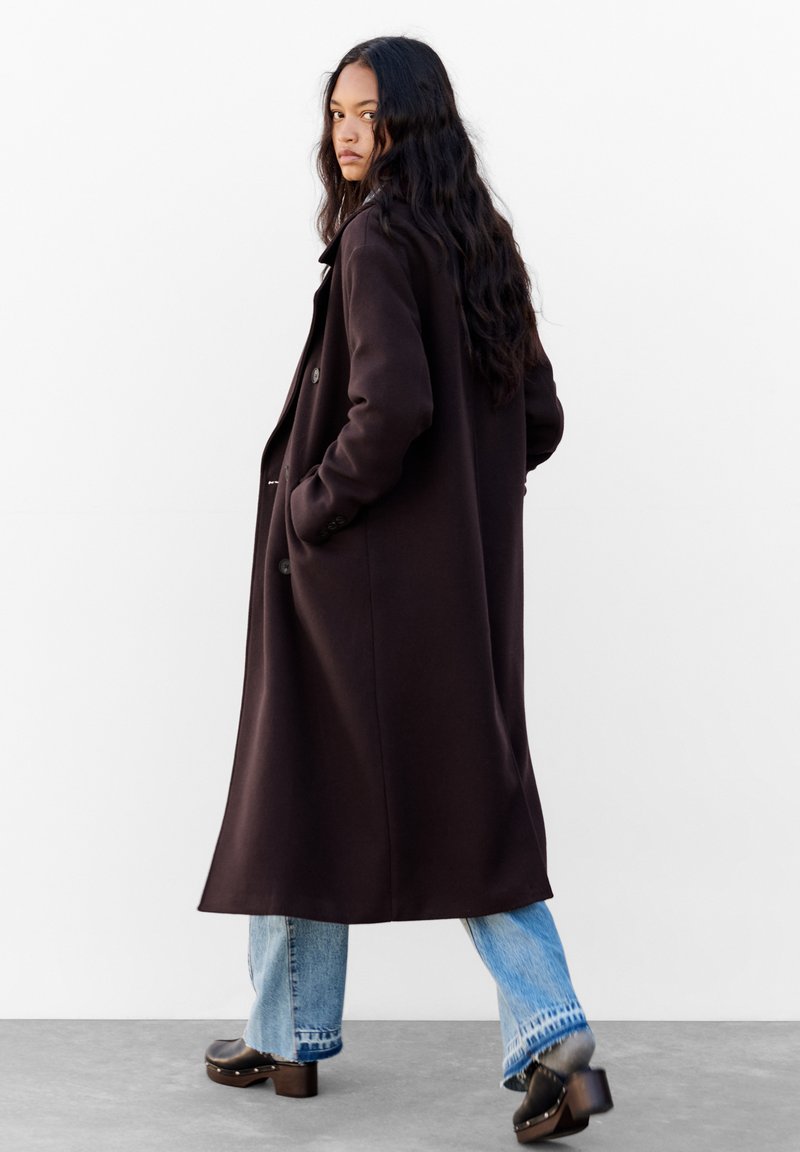 Długi, brązowy płaszcz z luźnym krojem, z guzikami, bocznymi kieszeniami i gładką teksturą. Połączony z jasnoniebieskimi dżinsami i butami na słupku.