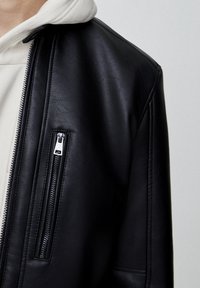 Svart läderjacka med dragkedja, slät textur och strukturerad design. Lager på en ljusfärgad hoodie.