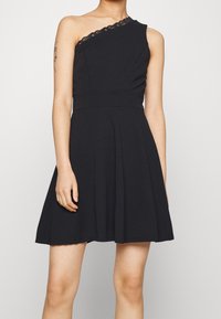 Černé šaty bez rukávů s jedním ramenem, zdobené krajkovými detaily kolem výstřihu a s rozšířeným designem sukně. Hladká textura látky.