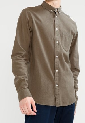 Mann trägt ein braunes, langärmliges Hemd mit Brusttasche und dunkelblaue Hose, steht vor einem einfarbigen Hintergrund.