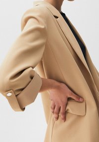 Beige blazer with a tailored fit, structured shoulders, and rolled cuffs. Features a side pocket and smooth, matte fabric texture.