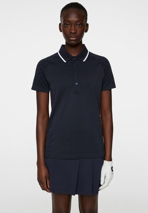 Polo shirt blu navy con colletto bianco e plastron con bottoni, dotato di pannelli laterali testurizzati. Abbinato a una gonna plissettata coordinata.